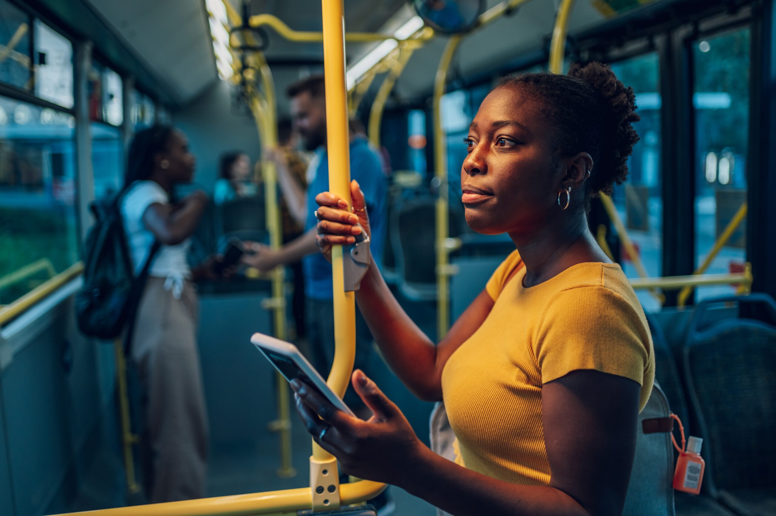 Young african american woman smiling while standing by herself on a bus and listening to music on a smartphone in the night. Female passenger using airpods and mobile phone in public transportation.